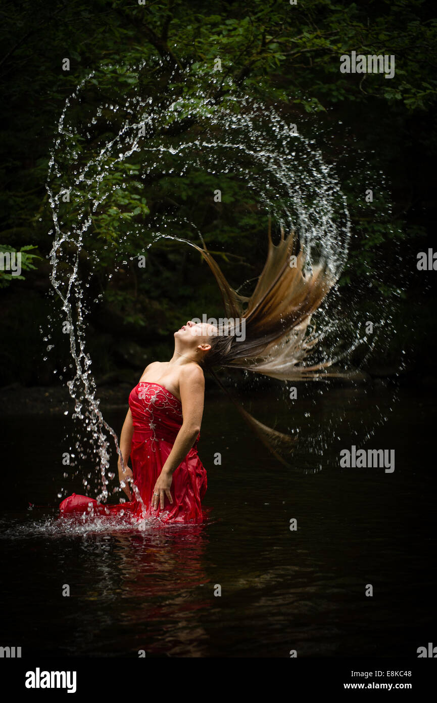 Sequenza di Fibonacci: una bionda dai capelli Ragazza donna che indossa un rosso intenso tonaca Palla abito in un fiume colpetti flipping i suoi capelli Foto Stock