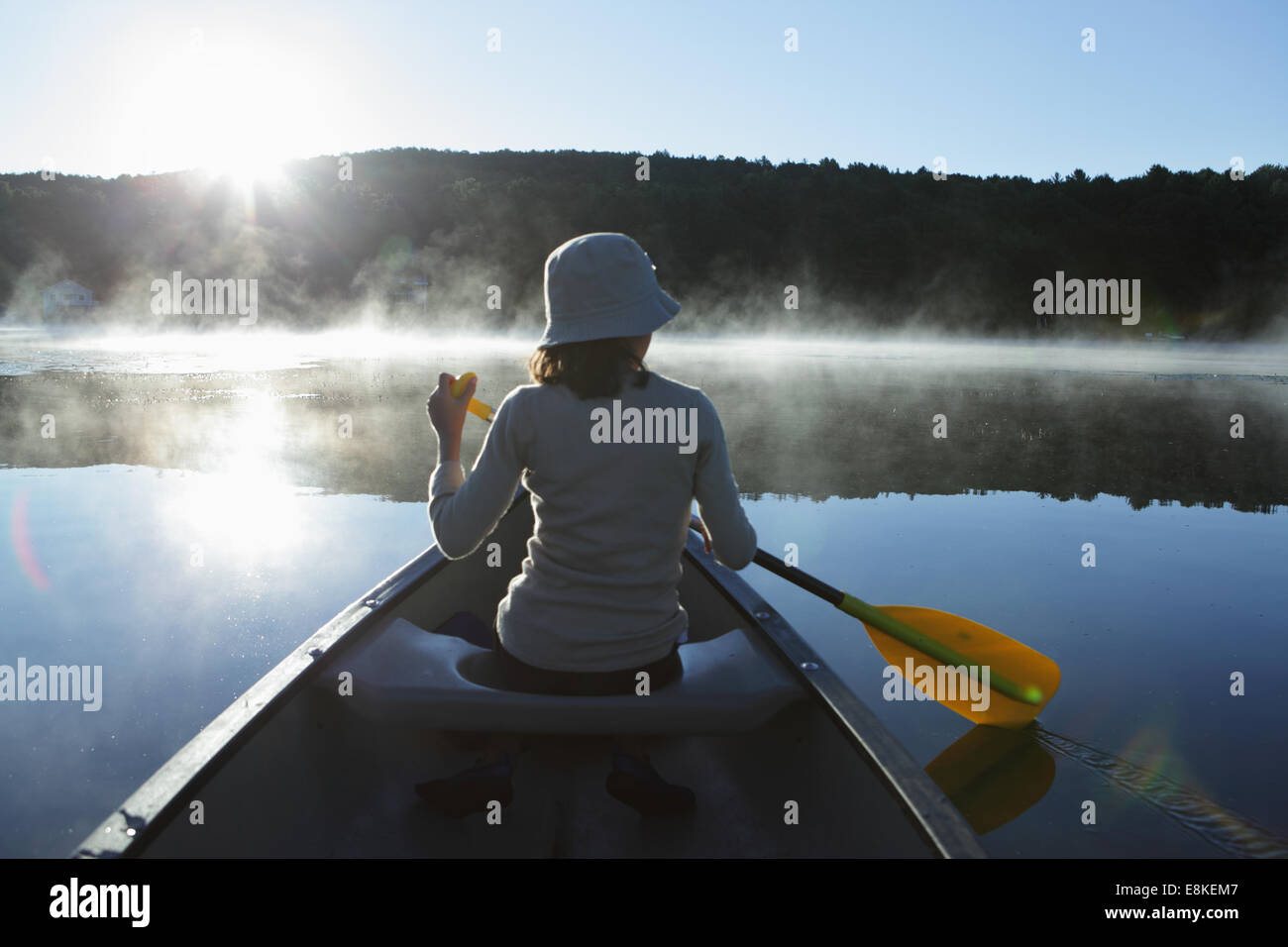 Ragazza giovane paddling una canoa in una nebbiosa lago calmo come il sole che sorge Foto Stock