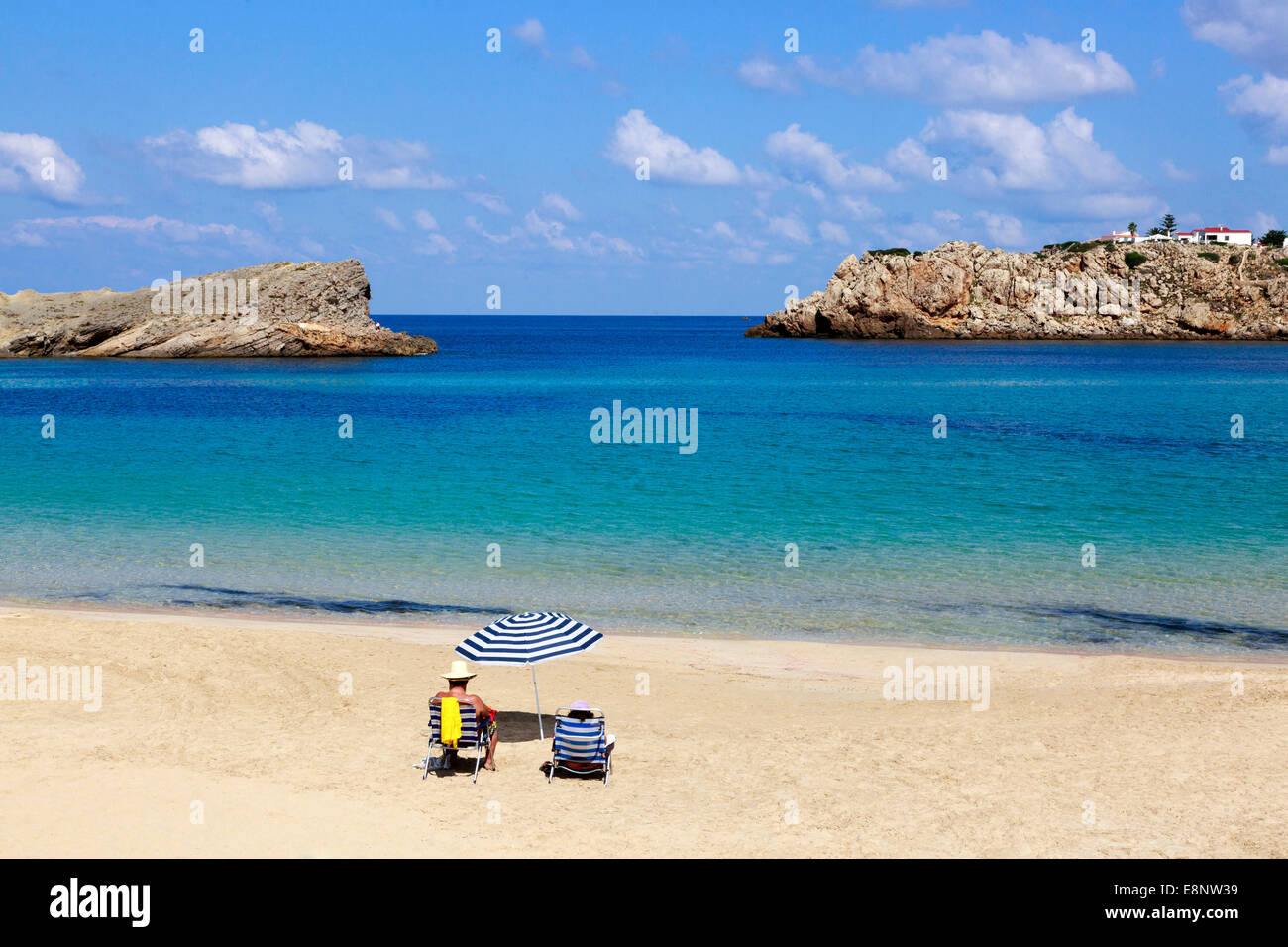 Uomo che indossa due cappelli, e una donna seduta sulla sdraio con un ombrellone, su una spiaggia in Arenel d'en Castell Foto Stock