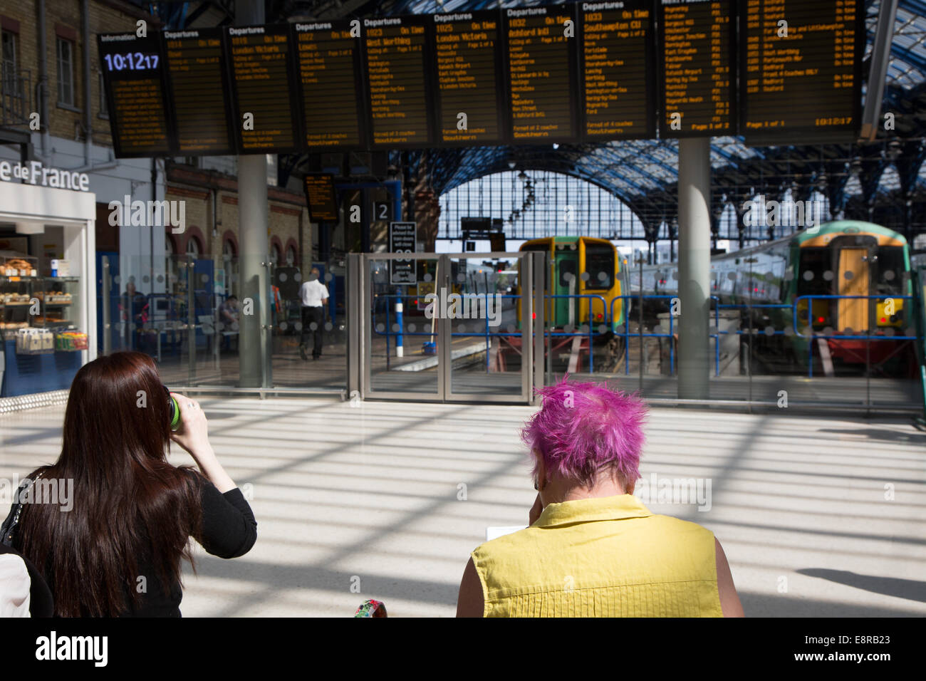 Attendere passeggeri per treno Informazioni sul piazzale presso la stazione del treno. Una delle signore ha carattere distintivo capelli rosa. Foto Stock
