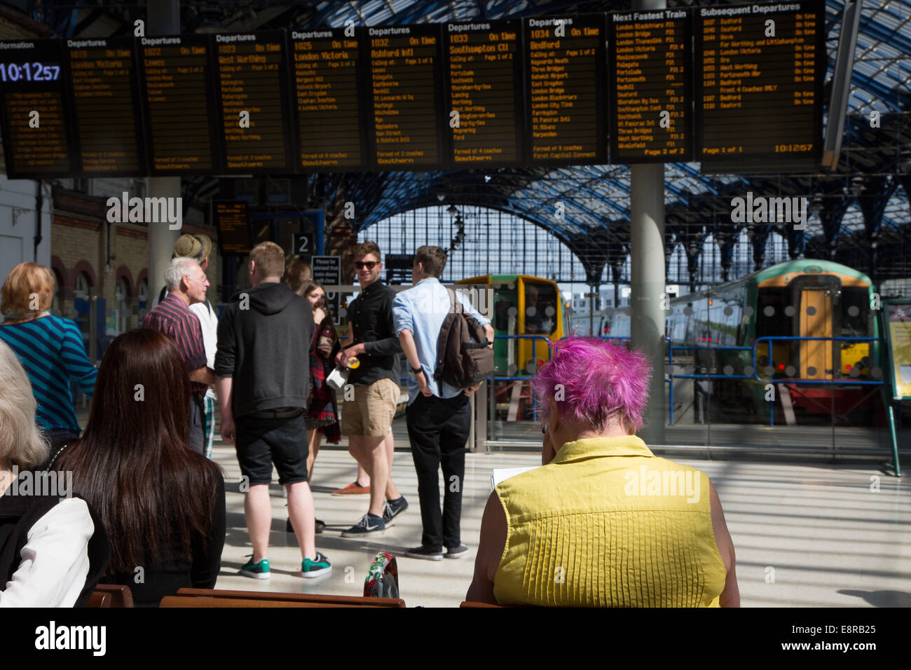 Attendere passeggeri per treno Informazioni sul piazzale presso la stazione del treno. Una delle signore ha carattere distintivo capelli rosa. Foto Stock