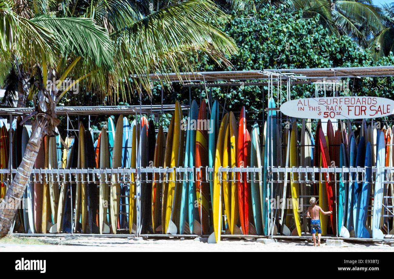 Un giovanissimo surfer guarda al classico degli anni sessanta le tavole da surf hawaiano che sono in vendita e in affitto sulla spiaggia di Waikiki a Honolulu sull'isola di Oahu nelle Hawaii, Stati Uniti d'America. Con il passare del tempo le tavole da surf sono evoluti da lunghe e pesanti tavole di legno degli antichi Hawaiiani a corto e leggero taglieri fatti di vari materiali come un nucleo schiumoso in poliuretano rivestito con tessuto in fibra di vetro che è rivestito con resina poliestere. Fotografato nel 1966. Foto Stock