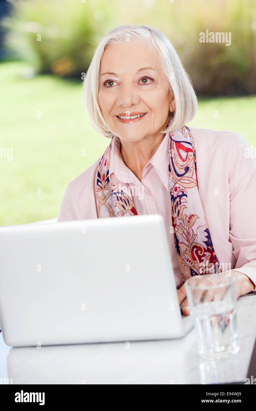 Sorridente donna anziana che guarda lontano mentre si utilizza sul computer portatile sul portico Foto Stock