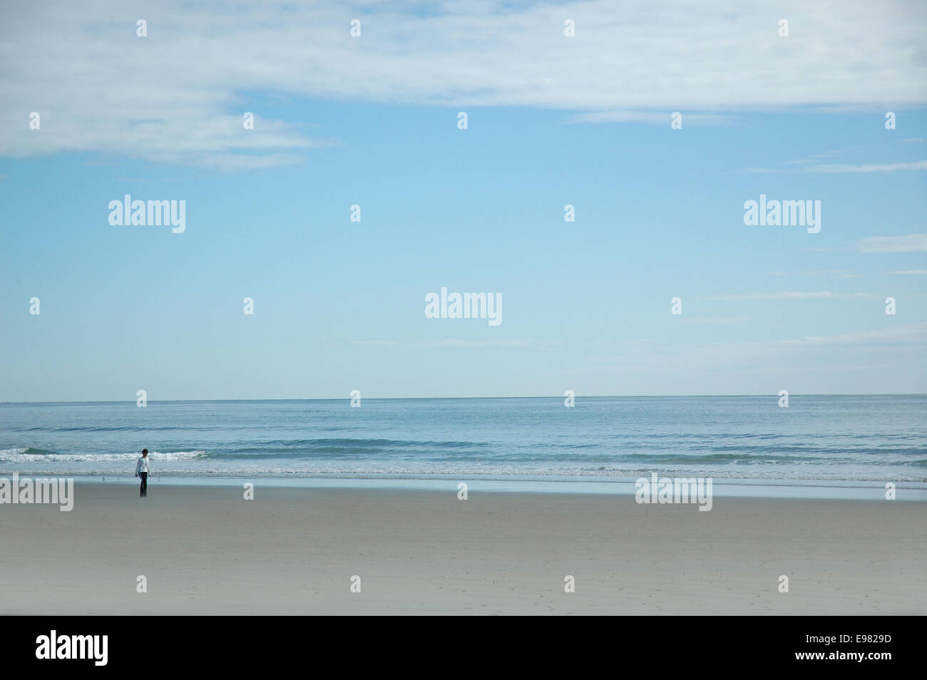 Donna adulta a camminare su una vasta distesa di spiaggia vicino al surf. Oceano e cielo sono di un colore turchese. Foto Stock