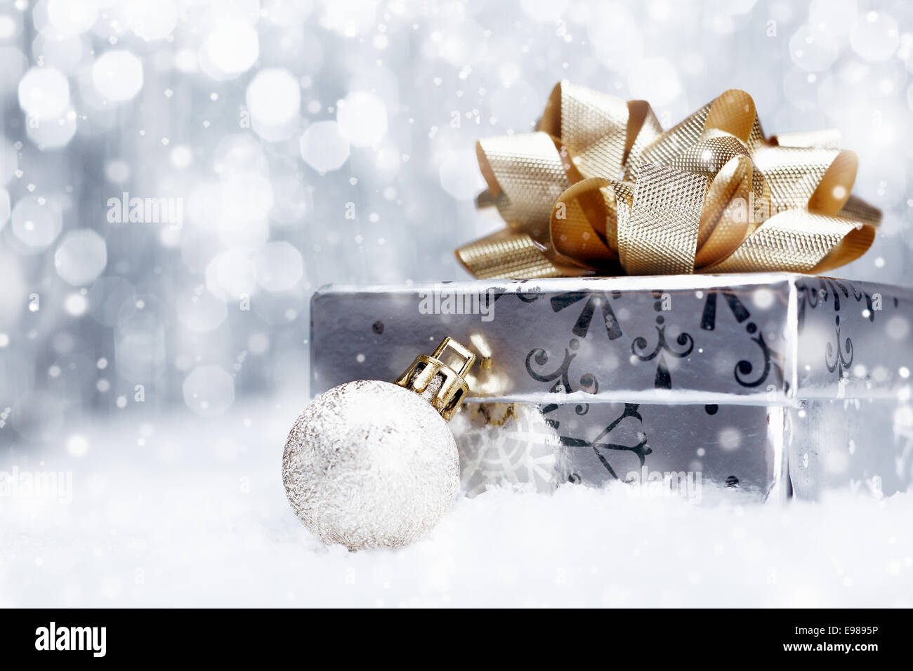 Argento regalo di Natale con un enorme ornamentali in golden bow giacente con la pallina su di un letto di neve fresca con la caduta di fiocchi di neve e copyspace Foto Stock