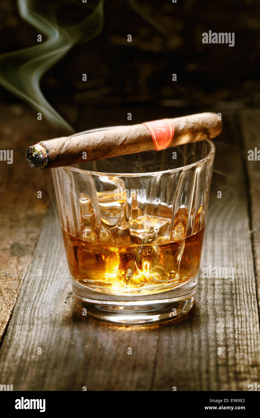 La masterizzazione di sigaro cubano in appoggio sul bicchiere di whisky su ghiaccio su un vecchio superficie in legno simbolica della mascolinità Foto Stock