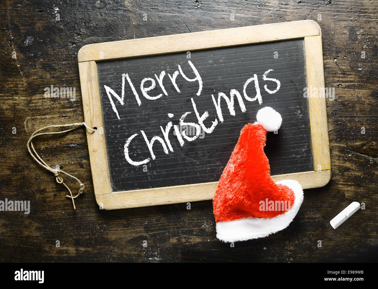 Manoscritti Merry Christmas saluto su una piccola lavagna con un lanuginoso rosso Santa hat su uno sfondo di legno Foto Stock