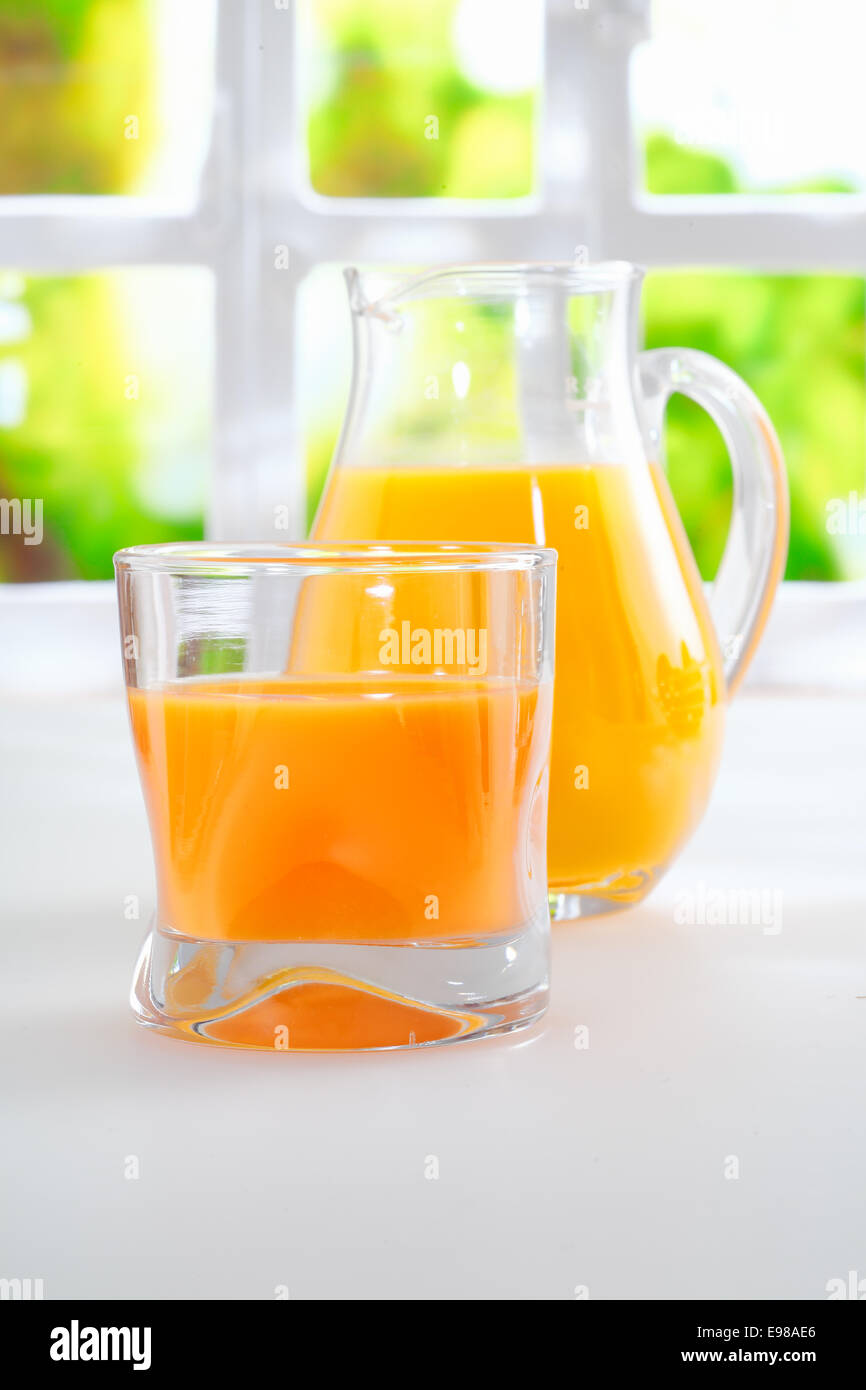Bicchiere di fresco e salutare il succo di arancia in piedi su un tavolo da cucina a fianco di una brocca piena di succo di frutta con una finestra piena di sole e di verde dietro Foto Stock