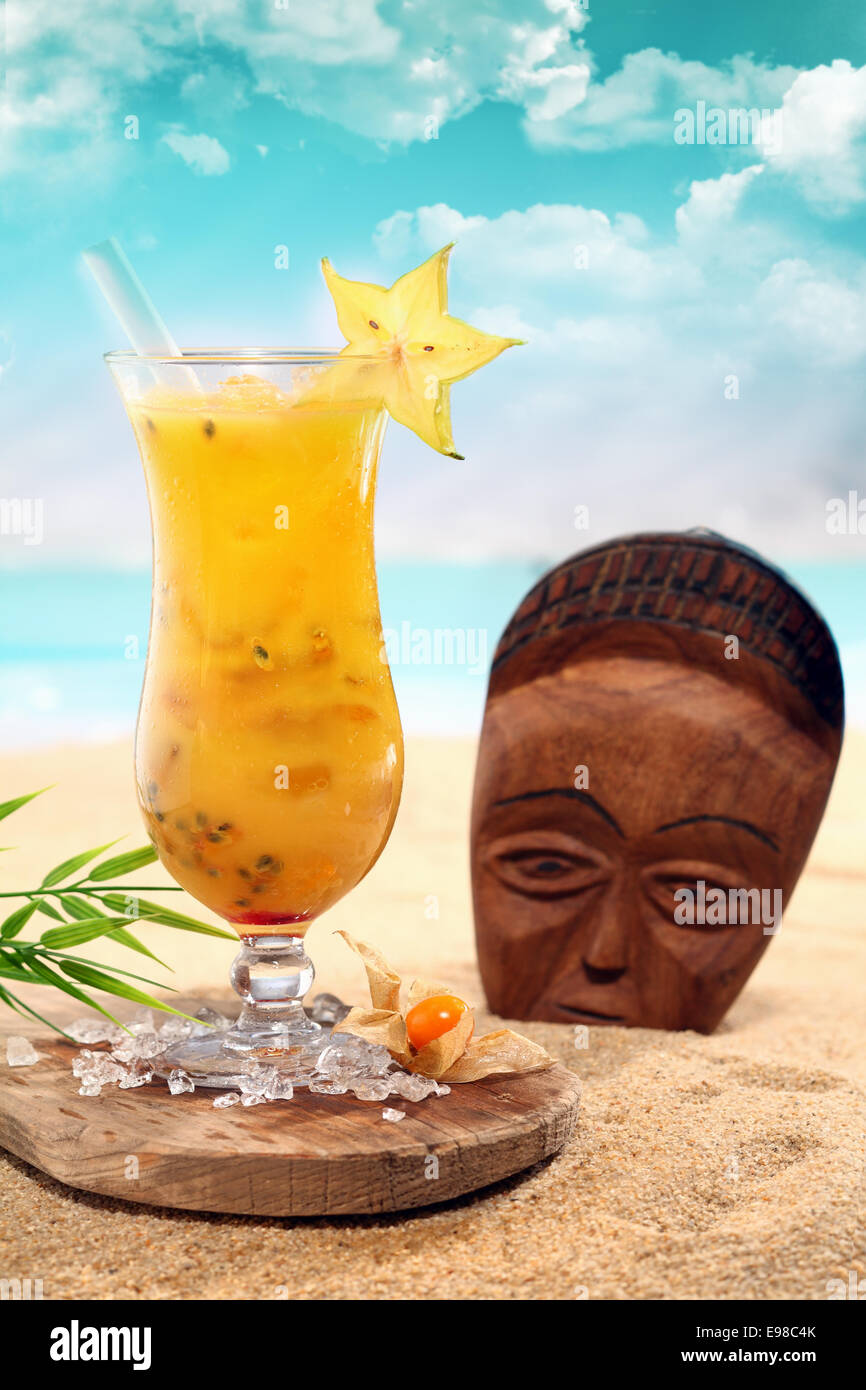 Colorato arancione ram ca bola cocktail in un alto eleganti vetri servita su un vassoio di legno sparsi con ghiaccio tritato a fianco di una maschera di legno ritti in spiaggia di sabbia su una spiaggia tropicale Foto Stock