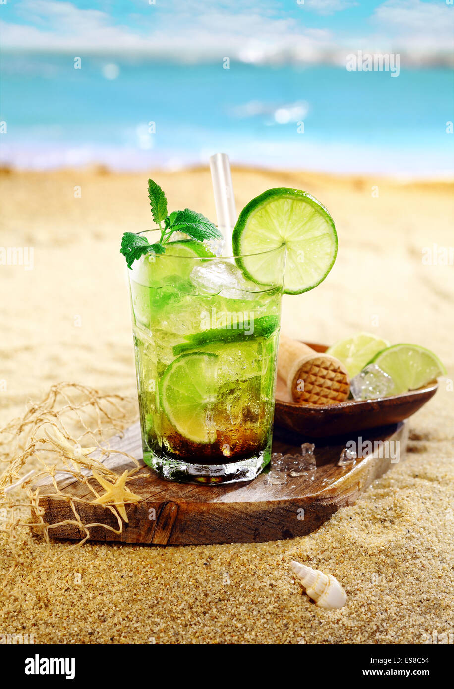 Caipirinha Rum e cocktail di calce guarnite con foglioline di menta e servita su una tavola di legno sulla sabbia dorata di una spiaggia tropicale Foto Stock