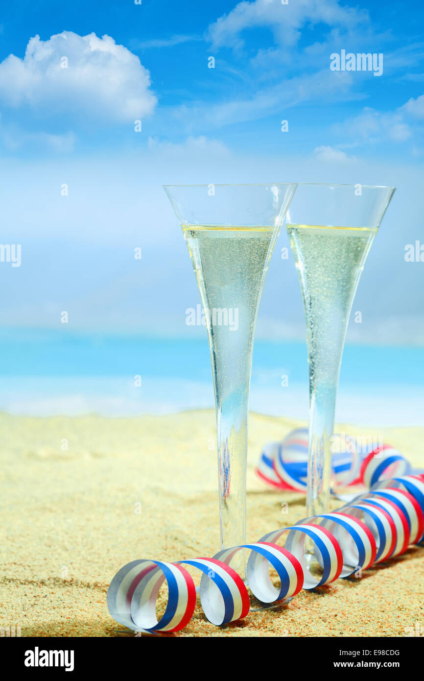 Champagne flauti in eleganti vetri scanalati e un colorato di rosso, bianco e blu partito twirled streamer permanente sulla sabbia dorata di una spiaggia tropicale con un blu azzurro Oceano su una vacanza estiva. Guardate il mio portafoglio per tutta una serie di cocktail. Foto Stock