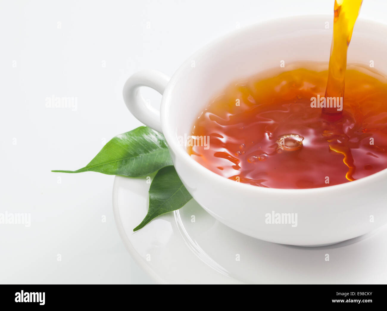 Vista ravvicinata di qualcuno versando una tazza di tè rinfrescante in una normale tazza bianca con fresco di foglie di tè verde in appoggio sul piattino su sfondo bianco Foto Stock