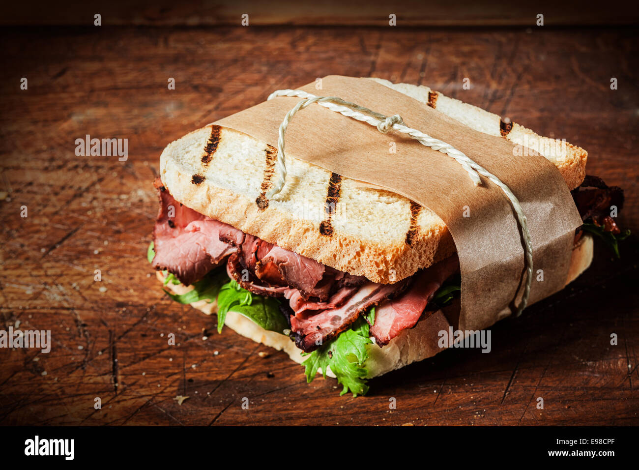 Deliziosa rare roast beef sandwich sul pane tostato alla brace avvolto in carta marrone legati con spago servita su un contatore di legno in un bistro in stile rustico Foto Stock