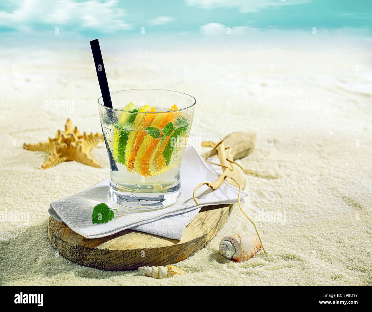 Bicchiere di rinfrescante o gin vodka cocktail con fettine di limone, calce e arancione su una spiaggia tropicale con un guscio e starfish sulla sabbia dorata Foto Stock