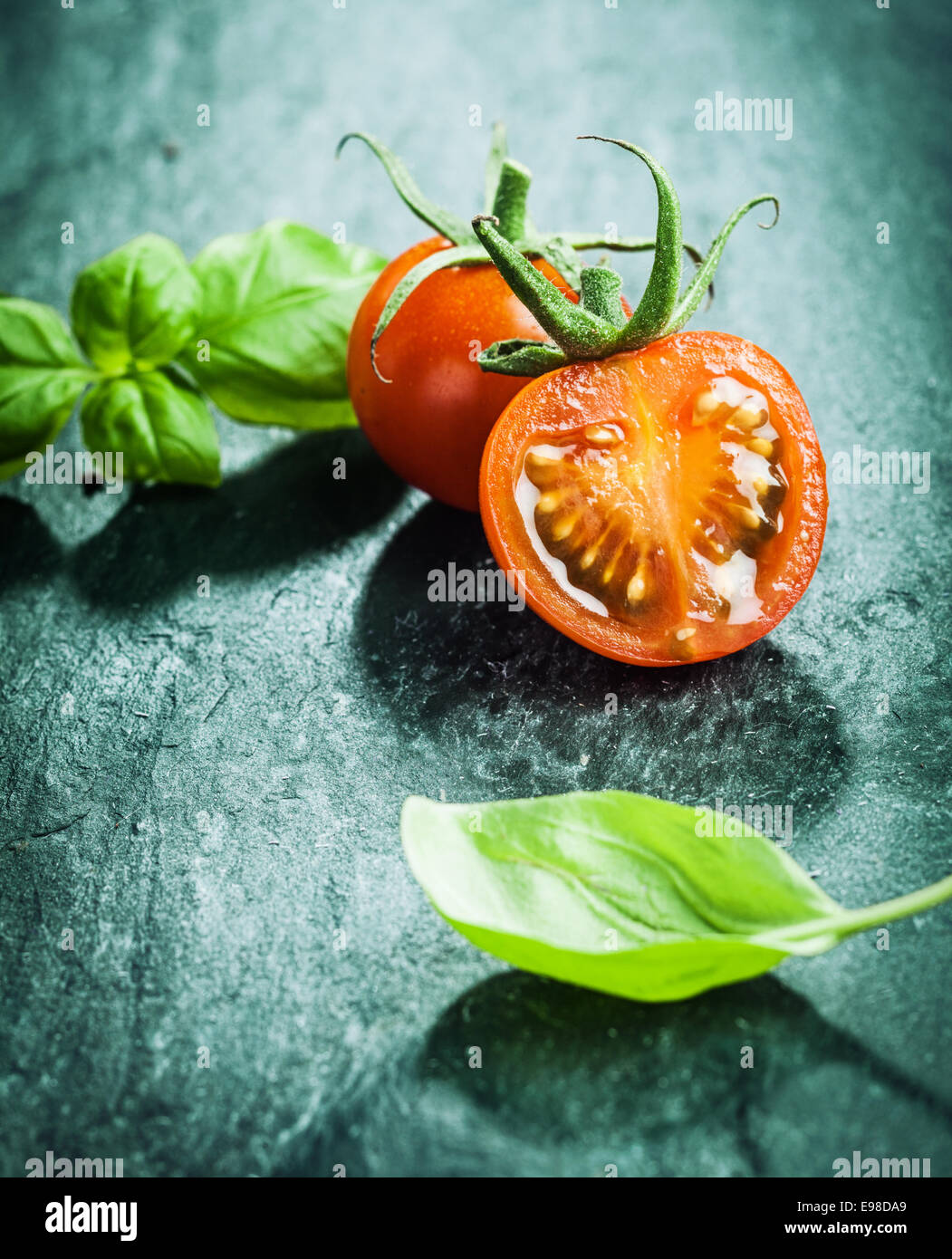 Di foglie di basilico fresco con dimezzato succosa ripe rosso pomodori uva pronta per la cottura di una pietanza saporita al buio su un superficie testurizzata Foto Stock