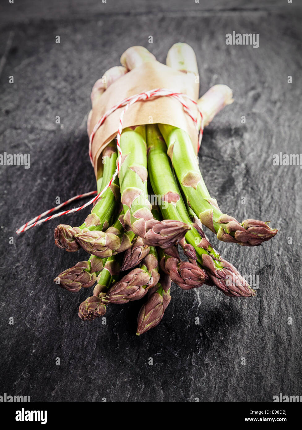 Freschi asparagi verdi spears raggruppati in carta marrone e legati con spago visualizzata con i loro suggerimenti per la telecamera su un grigio ardesia testurizzata superficie Foto Stock