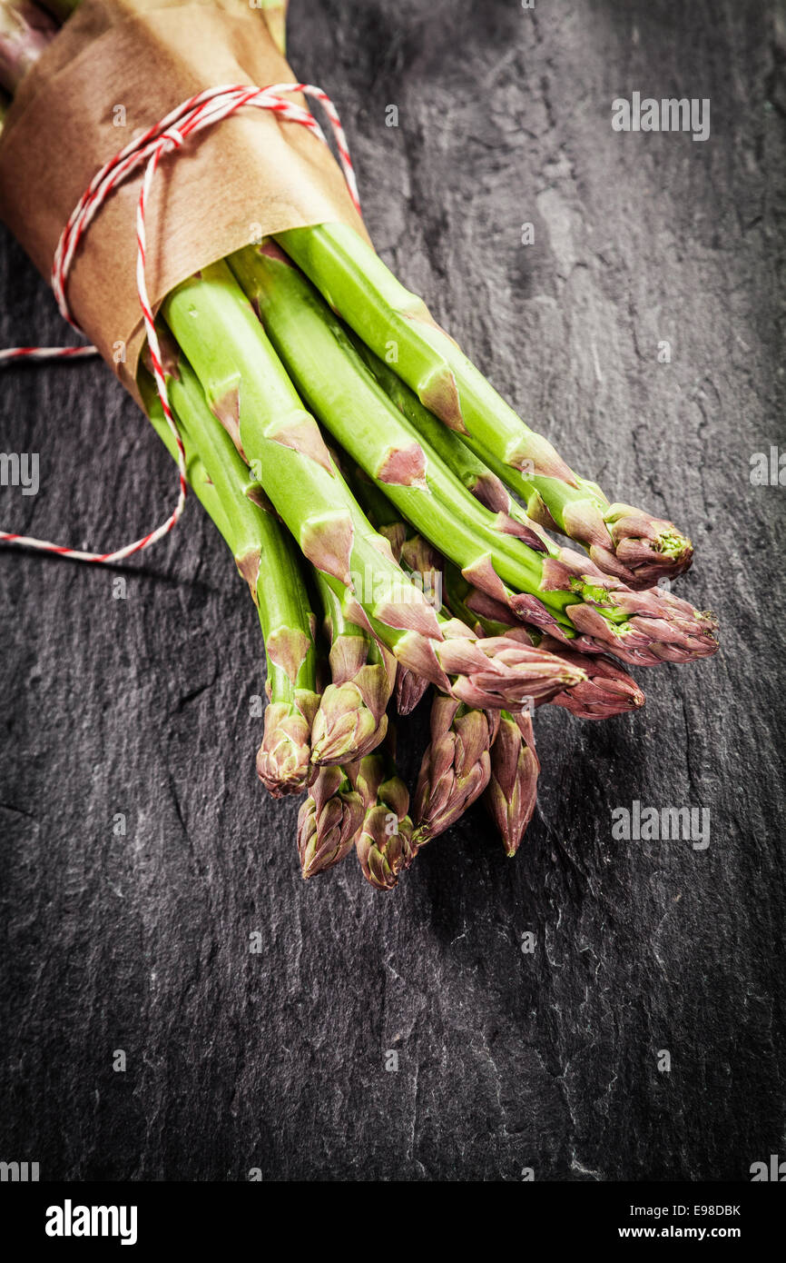 Fattoria fresco di punte di asparagi in un involucro marrone legati con spago giacente su una superficie strutturata di ardesia grigia con superficie di vignettatura e copyspace per il testo Foto Stock