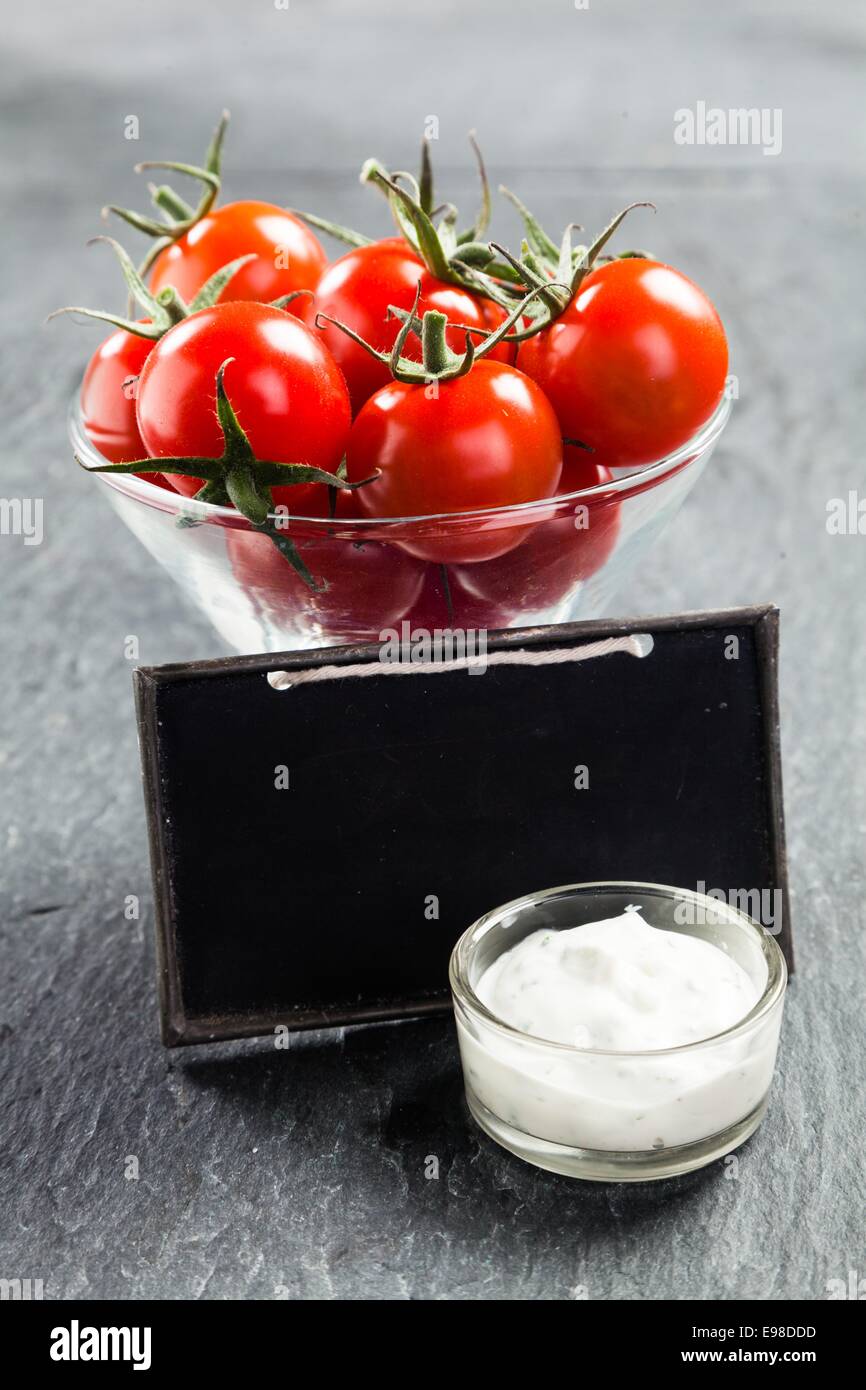 Ciotola di vetro di freschi maturi rossi pomodori ciliegini e saporita cremosa salsa vinaigrette su un sfondo di ardesia con un vuoto scuola vintage ardesia con copyspace per il testo Foto Stock