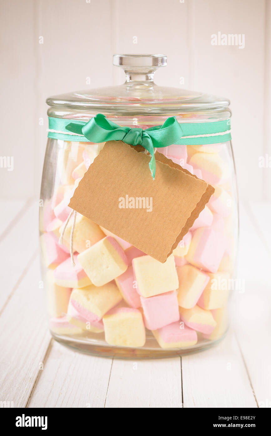 Vetro Antico caramelle vaso riempito con quadrato rosa e giallo marshmallows e un tag vuoto legate con un nastro di verde su un bianco sullo sfondo di legno Foto Stock
