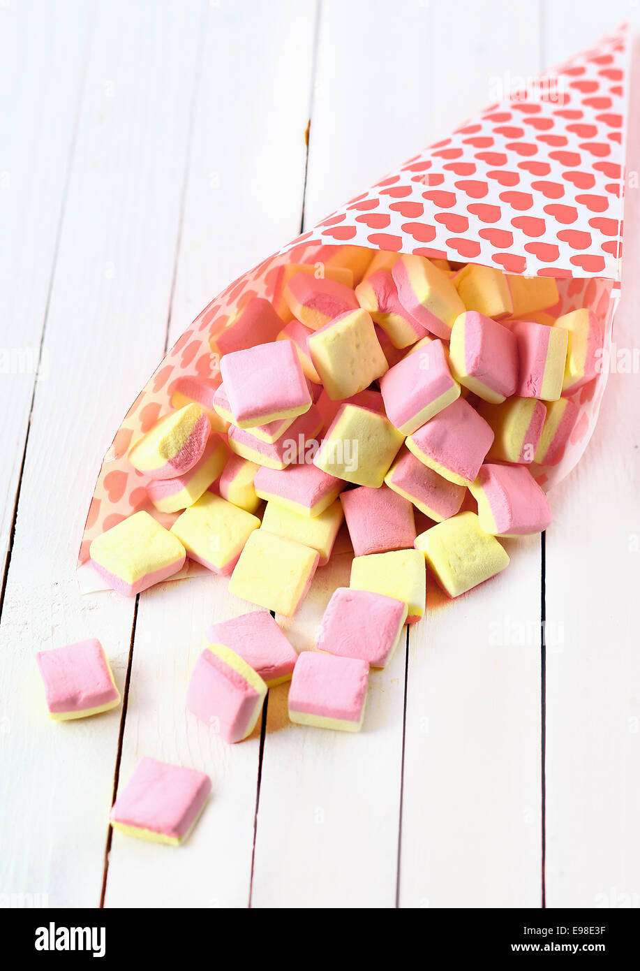 Vista ravvicinata di un cono di caramella decorata con cuori rosa fuoriuscita di forma quadrata rosa e giallo marshmallows oltre un legno bianco bordo tabella Foto Stock