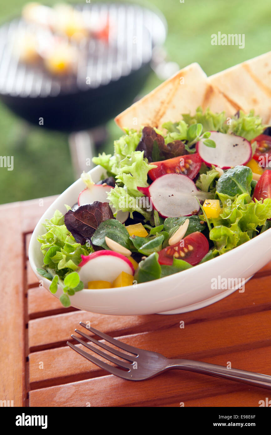 Veggie sani e freschi verde mista insalata vegetariana su una tavola di legno tavolo da picnic con fette di arrosto di focacce e un barbecue estivo visibile sul prato al di là Foto Stock