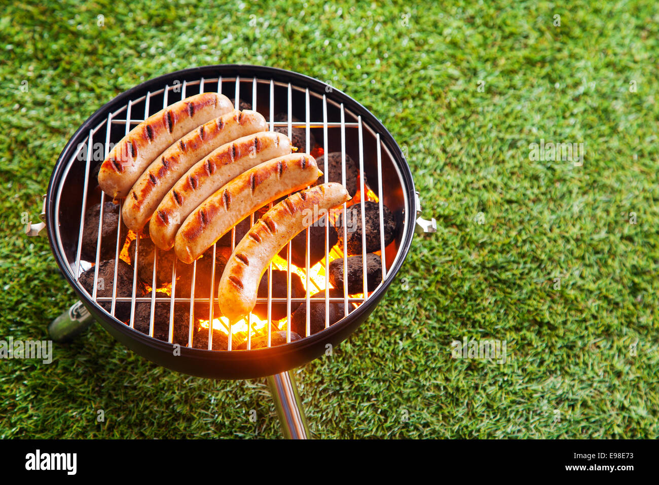Grigliare le salsicce oltre il caldo carboni ardenti in un barbecue portatile in piedi su un prato verde con copyspace durante un picnic o Estate viaggio di campeggio Foto Stock