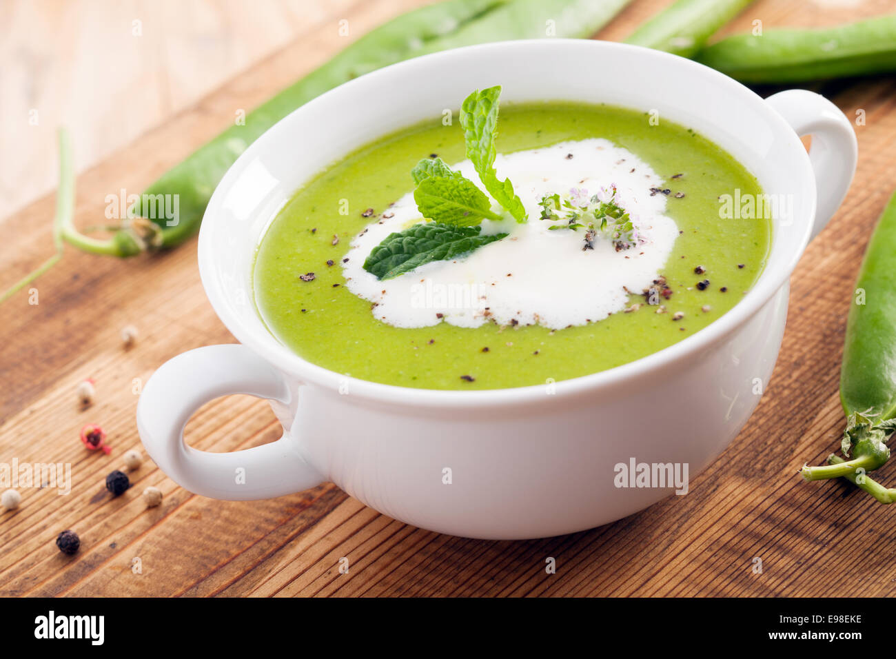 Close up di una ciotola di crema verde di zuppa di piselli guarnita con panna e erbe aromatiche fresche su una superficie in legno con sparse baccelli di piselli Foto Stock