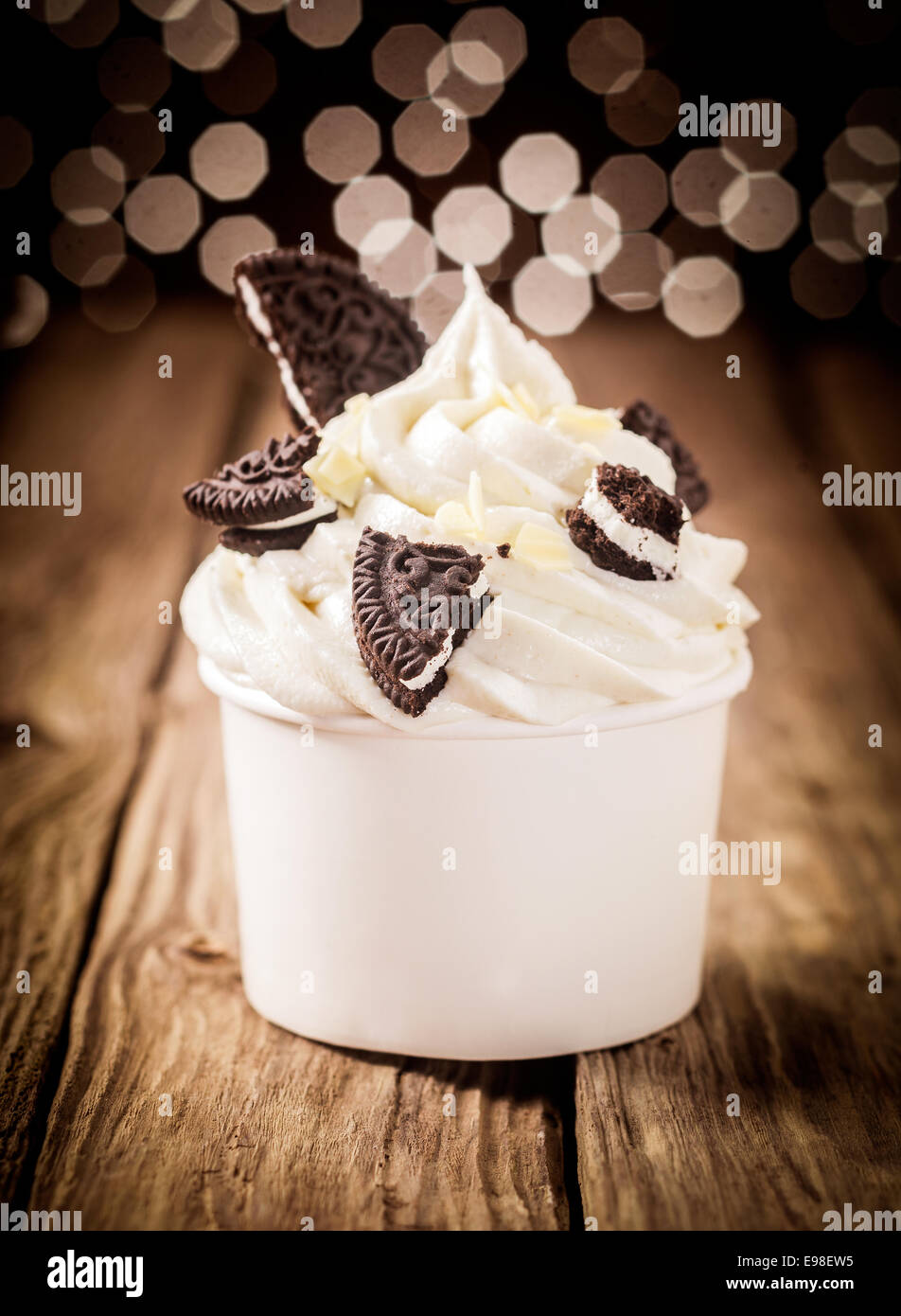 Pezzi di cioccolato rotto oreos su un mulinare di ricca crema di yogurt surgelato in una plastica vasca da asporto su un banco di legno con un bokeh di luci scintillanti Foto Stock