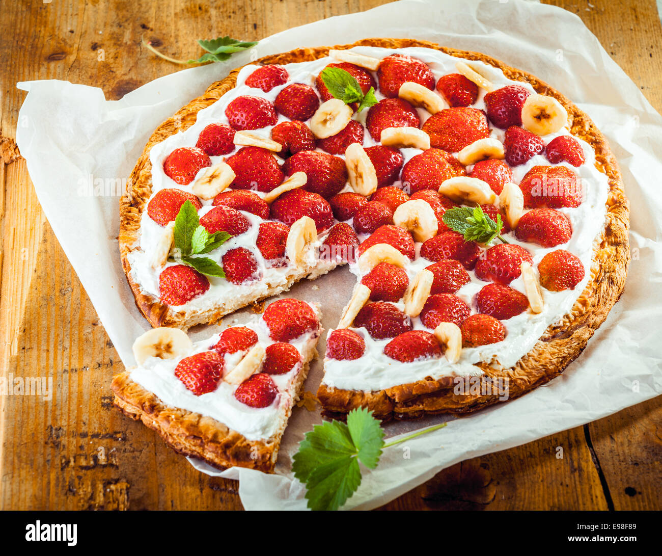 Appena sfornati e fragola flan di banana con un croccante crosta dorata e topping di frutti di bosco freschi , di banana e panna guarnita con la menta piperita su un quadrato di carta bianca, ad alto angolo di visione Foto Stock