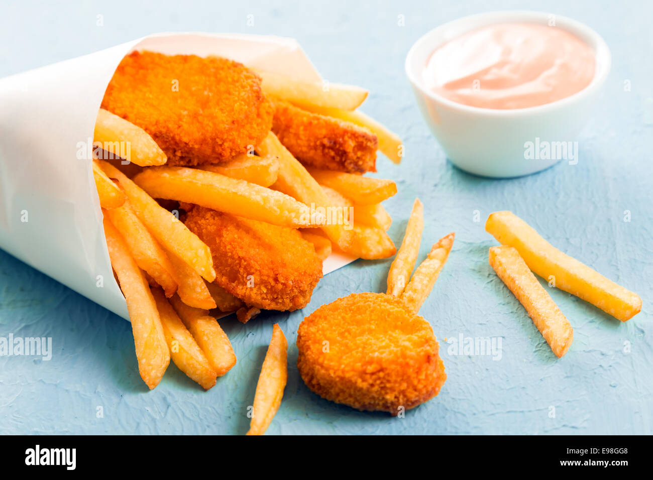 Pesce fritto pepite d'oro con le patatine fritte fuoriusciti da un cono di carta da un pesce da asporto shop su una superficie di colore blu, vista ravvicinata Foto Stock