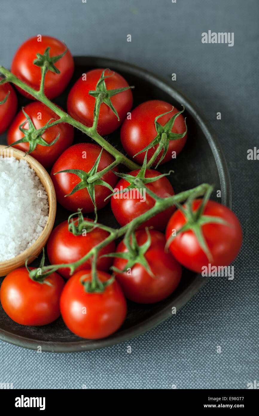 Mazzetto di ripe rosso pomodori ciliegia sulla vite disposti su una piastra con una ciotola di grossolana del sale marino naturale per uso come cottura Ingredienti in cucina Foto Stock