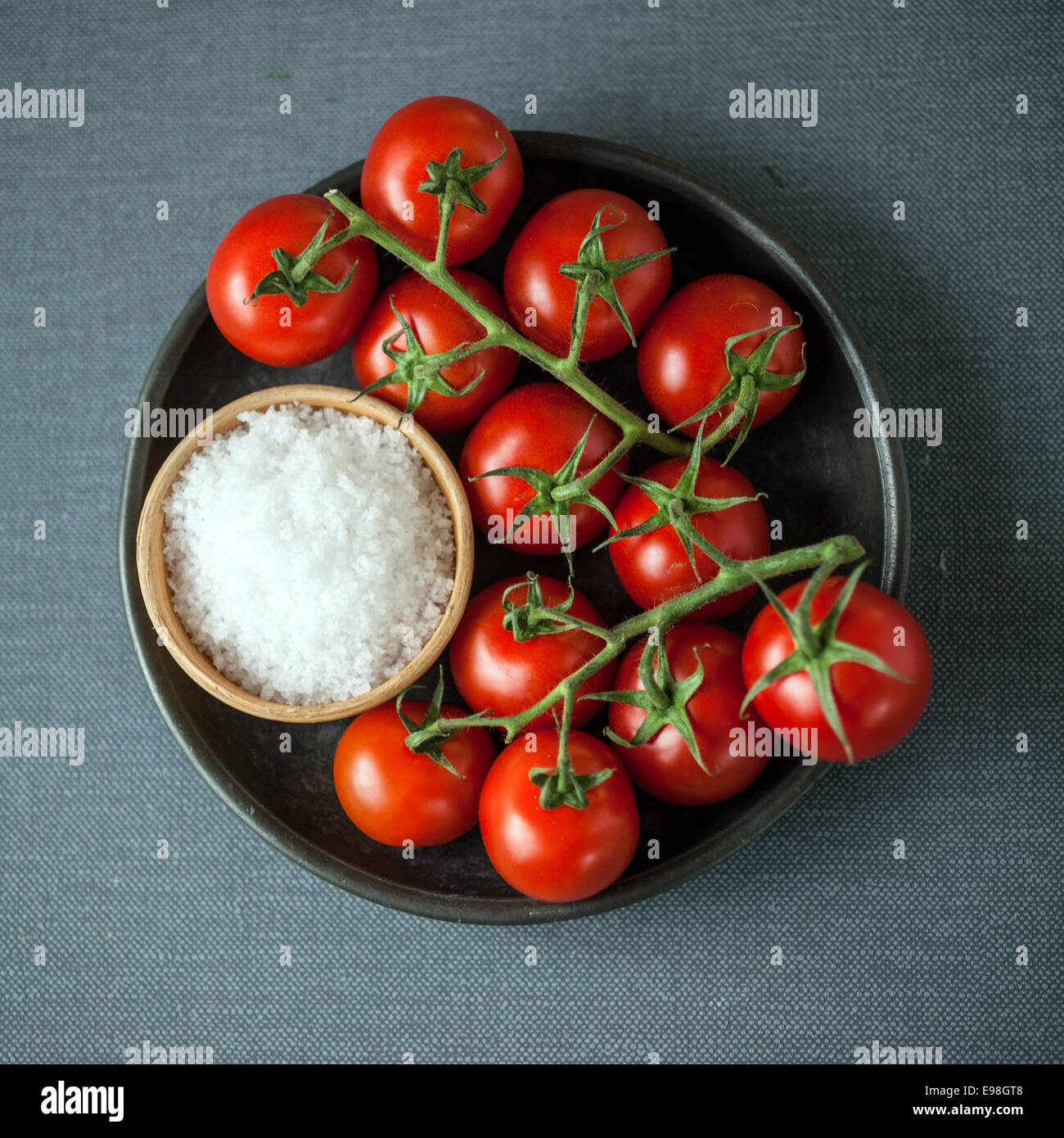 Vista aerea di un mazzetto di ripe rosso pomodori ciliegia sulla vite e una ciotola di grossolana del sale marino naturale per la cottura Ingredienti su un tessuto grigio Sfondo in formato quadrato Foto Stock