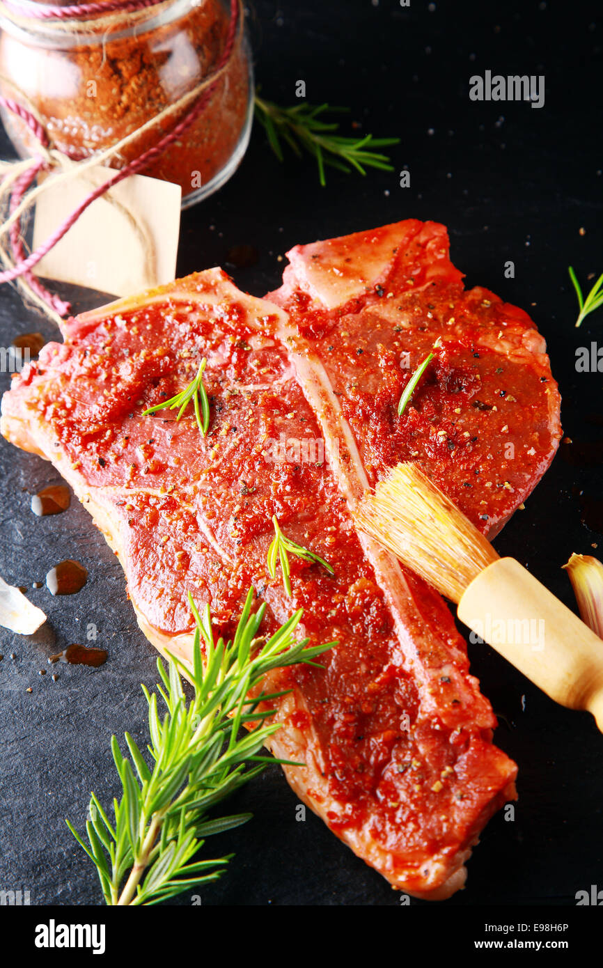 Materie osteria bistecca con condimento piccante e un rametto di rosmarino fresco a fianco di un vasetto di spezie in polvere con un'etichetta vuota durante la preparazione della carne per la griglia in una cucina rustica Foto Stock