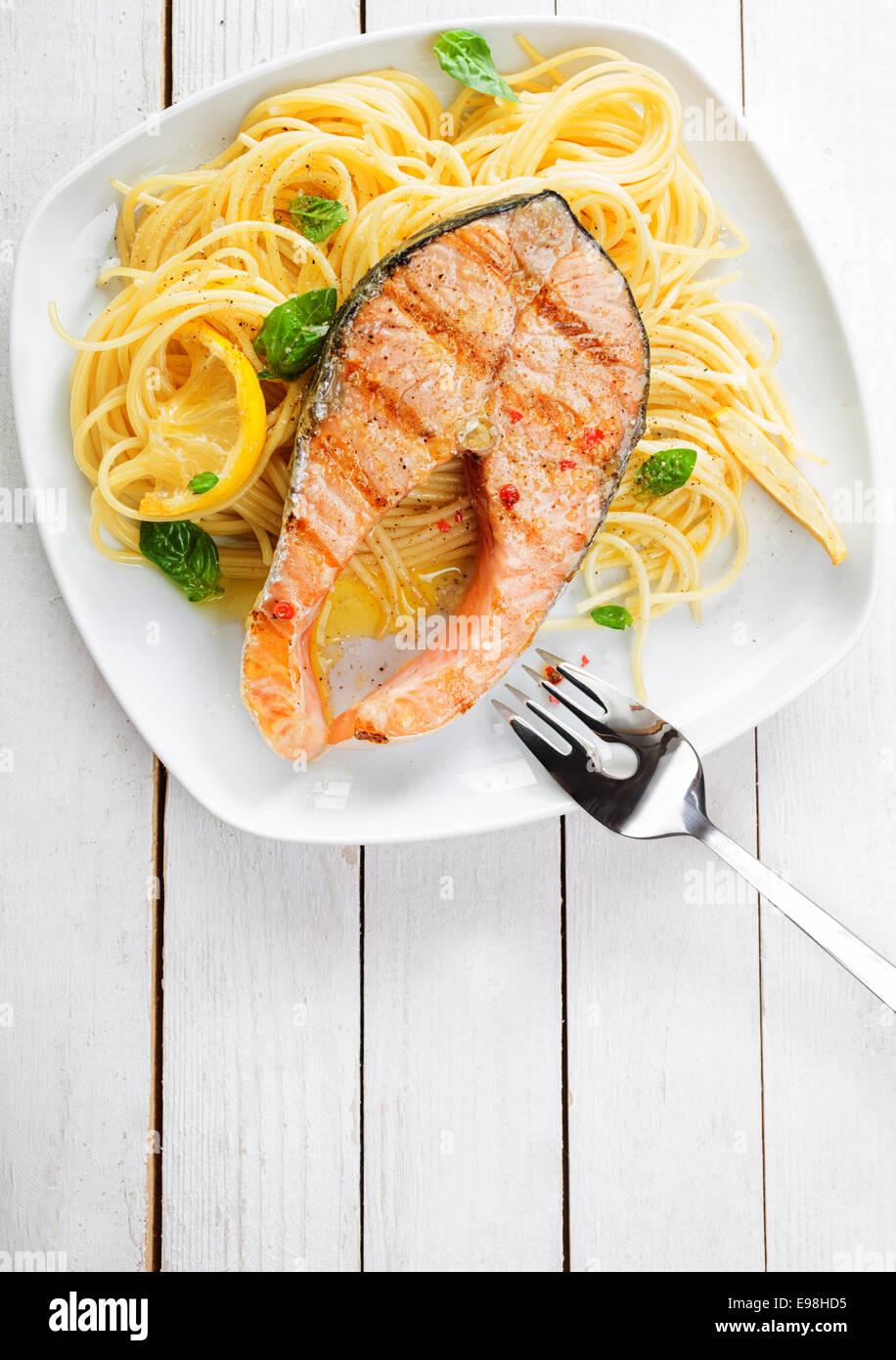 Gourmet cucina a base di pesce Con Salmone alla griglia cotoletta bistecche servite su un letto di italiano linguine guarnita con limone e basilico fresco, vista aerea su un bianco tavolo in legno con copyspace Foto Stock