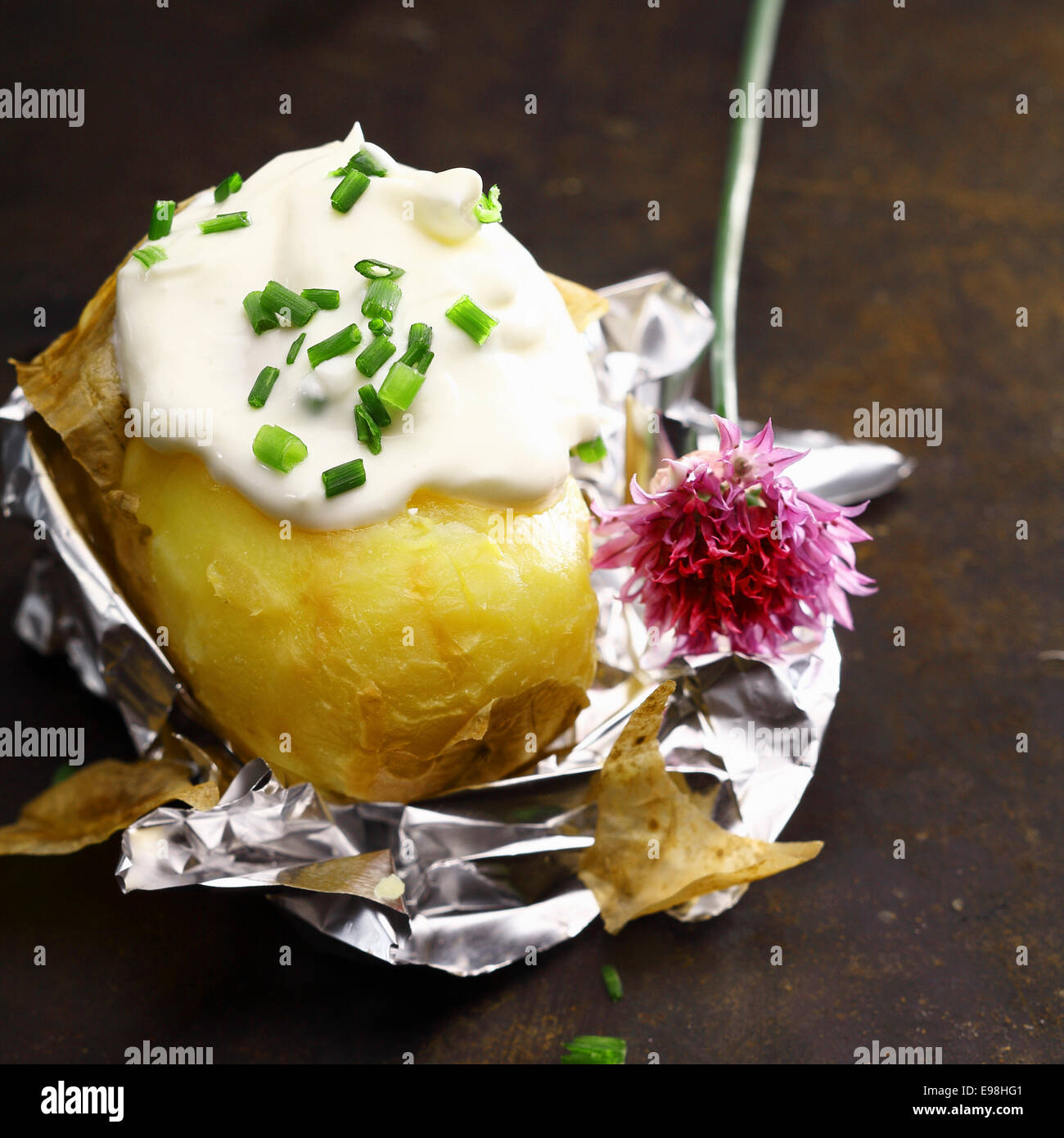 Lamina di patate al forno con panna acida e erba cipollina fresca tritata con un fiore fuori da una pianta di erba cipollina servita parzialmente pelato sul suo involucro in foglio Foto Stock