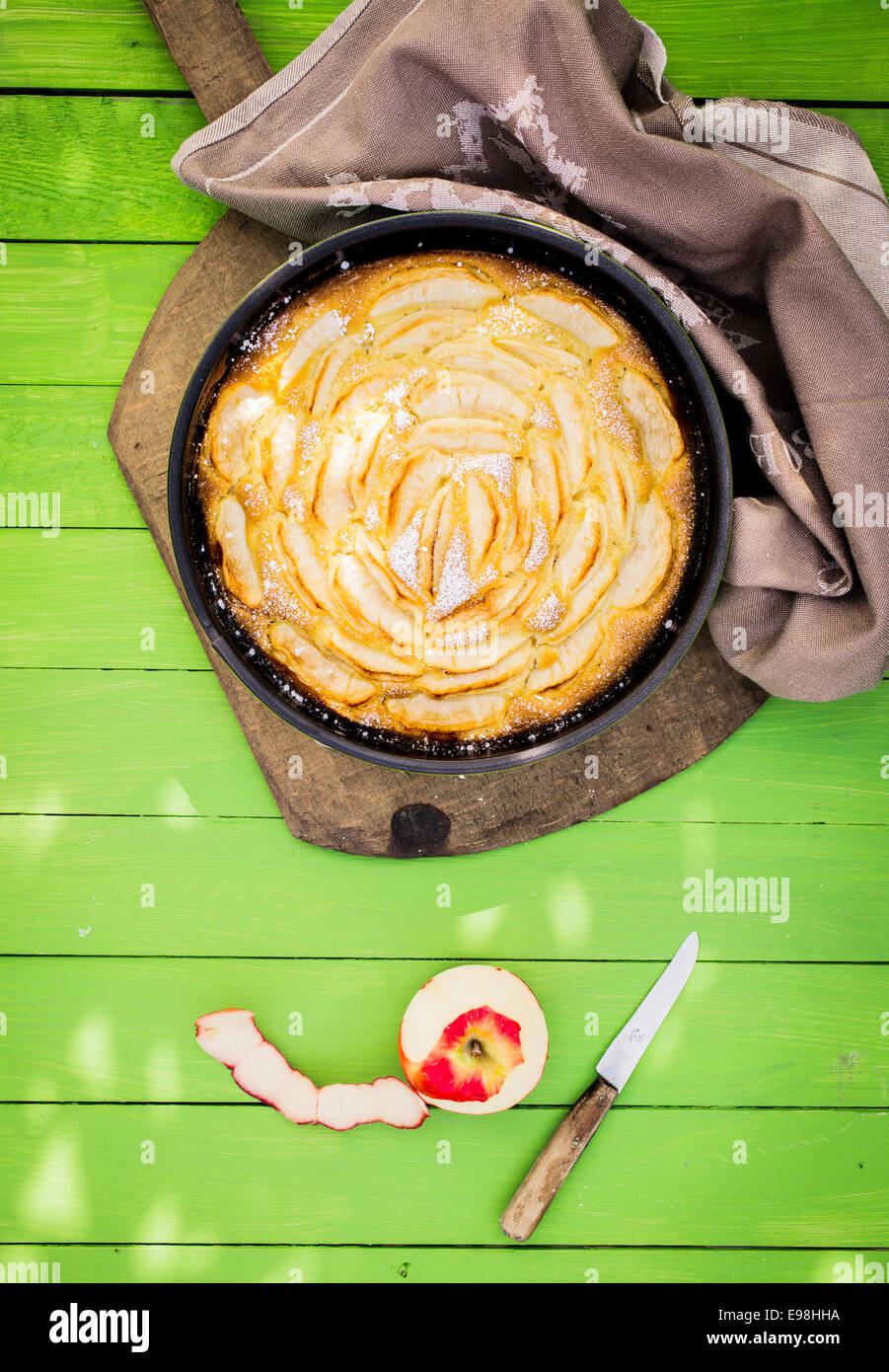 Fatti in casa crostata di mele cotte con mele fresche formano il giardino servita su un verde giardino in legno tavolo in pezzata di luce solare con una mezza mela sbucciata e coltello seghettato a fianco Foto Stock
