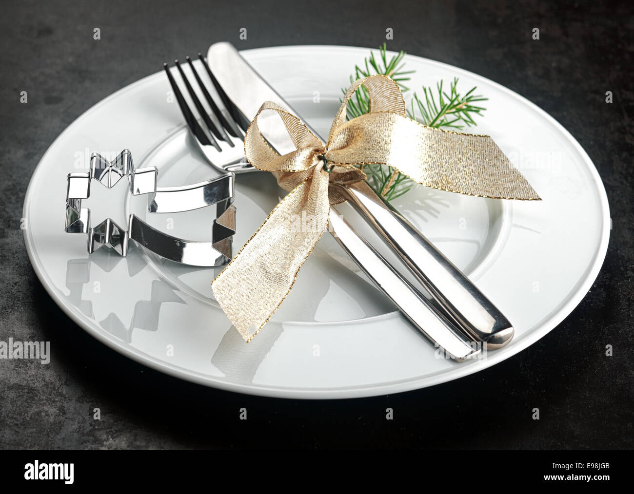 Festa di Natale posto impostazione con un argento lucido shooting star cookie cutter a fianco di un coltello e forchetta legato decorativa con un nastro color oro con un ciuffo di fronde di pini su una pianura piastra bianca Foto Stock