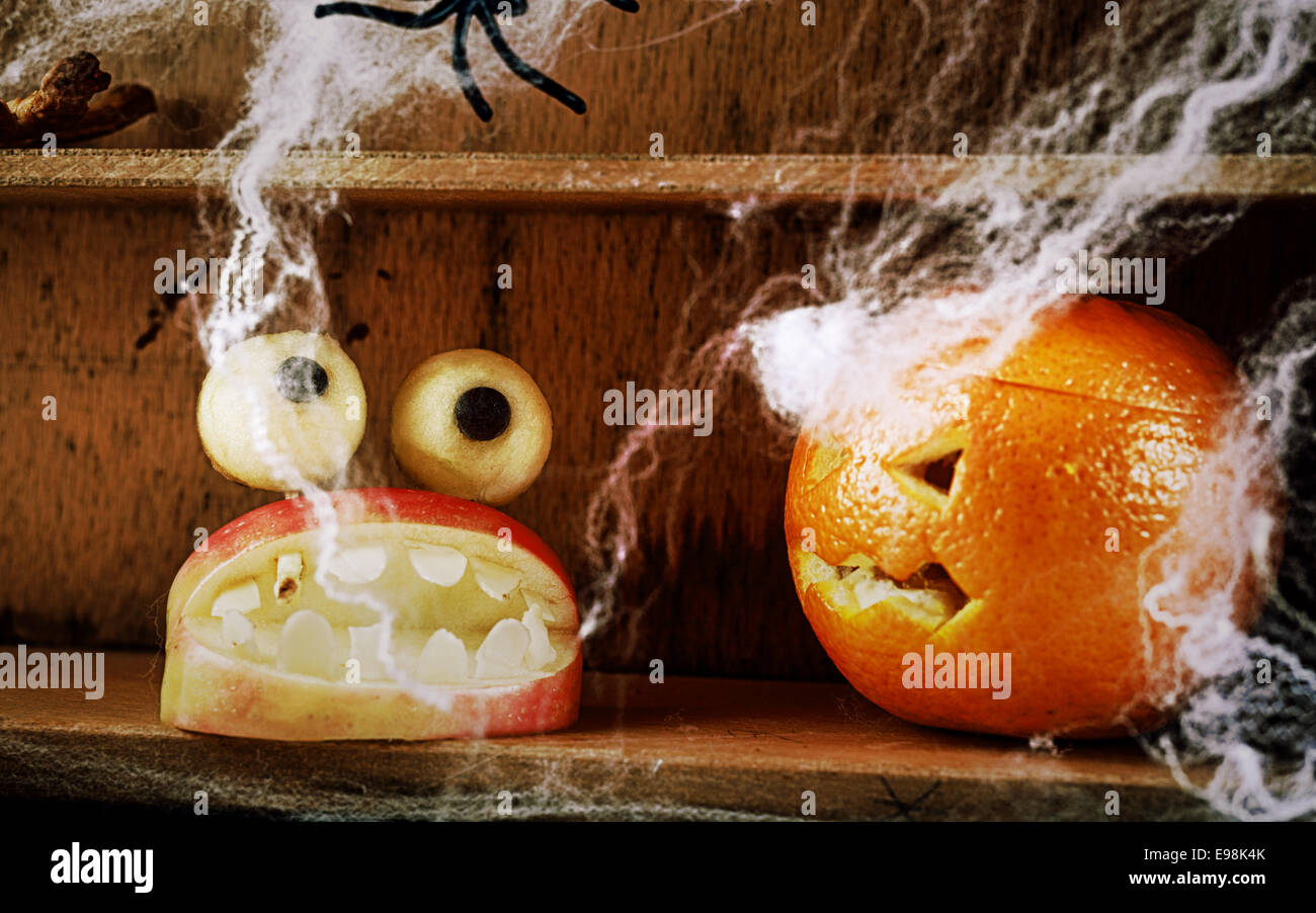 Divertimento in casa spooky Halloween cibo decorazioni con un Apple intagliato bocca piena di temibili denti e rabboccato con impasto rotondo occhi e un jack-o-lantern arancione scolpito su un ripiano di legno con ragni e nastri Foto Stock