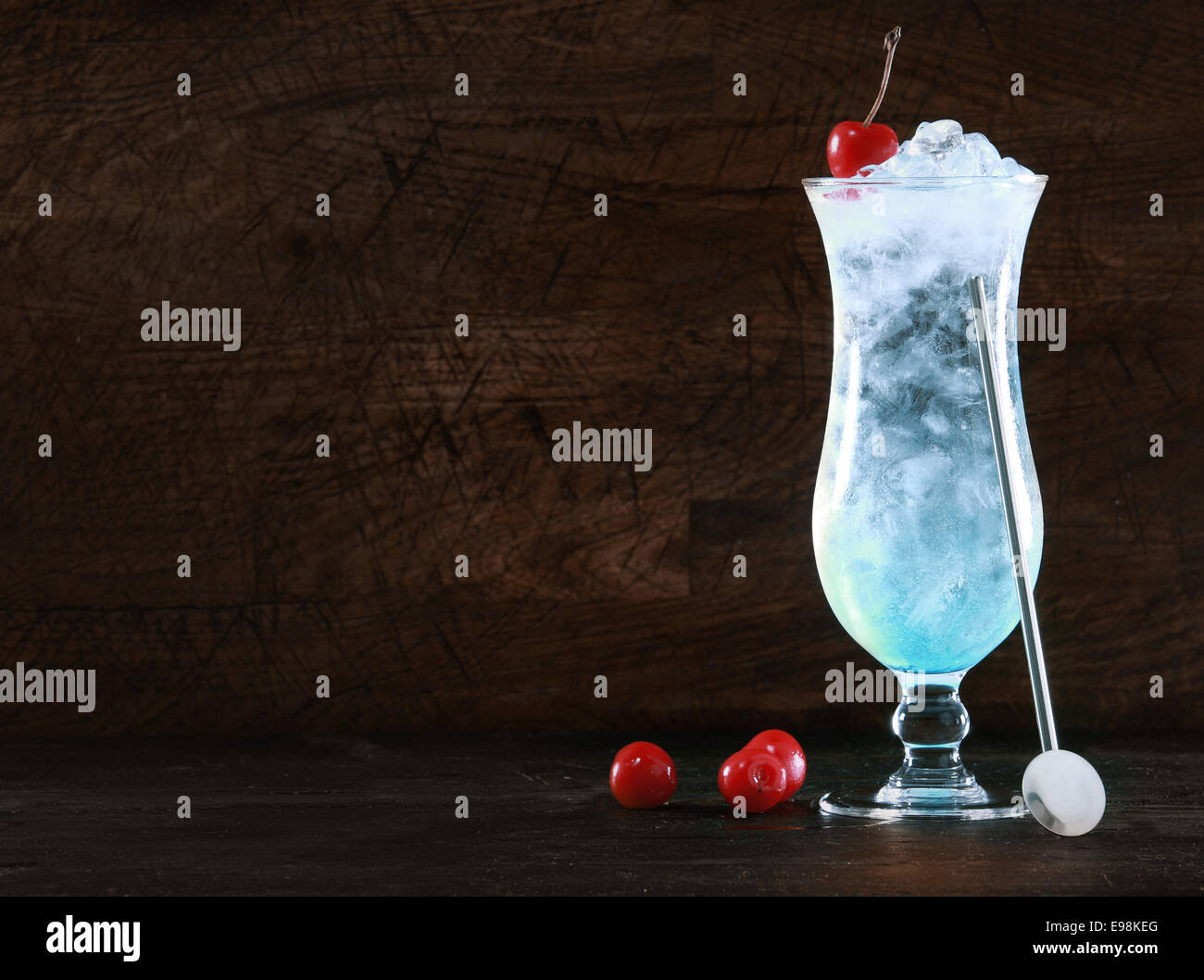 Elegante curacao blu cocktail con ghiaccio e un swizzle-stick guarnita con ciliegie fresche su sfondo scuro con copyspace per il tuo messaggio di Natale o di vacanza tropicale testo Foto Stock