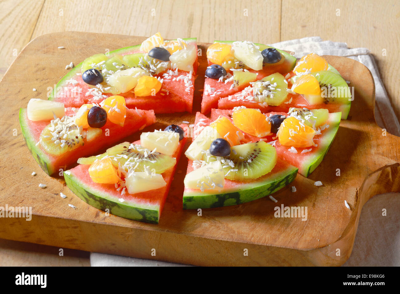 Basso angolo di visione di un sano vegetariano e vegano frutta tropicale anguria pizza guarnita con kiwi, mirtilli, arancia, ananas, e cosparso di cocco essiccato, tagliato in segmenti per servire Foto Stock