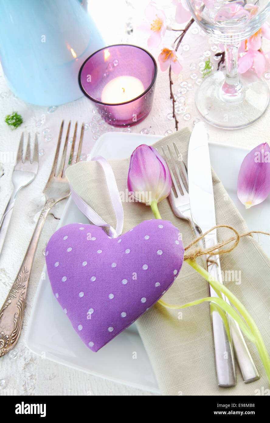 Elegante luogo romantico impostazione con un cuore lilla per un sweetheart circondato da argenteria, una candela e delicati fiori di primavera Foto Stock