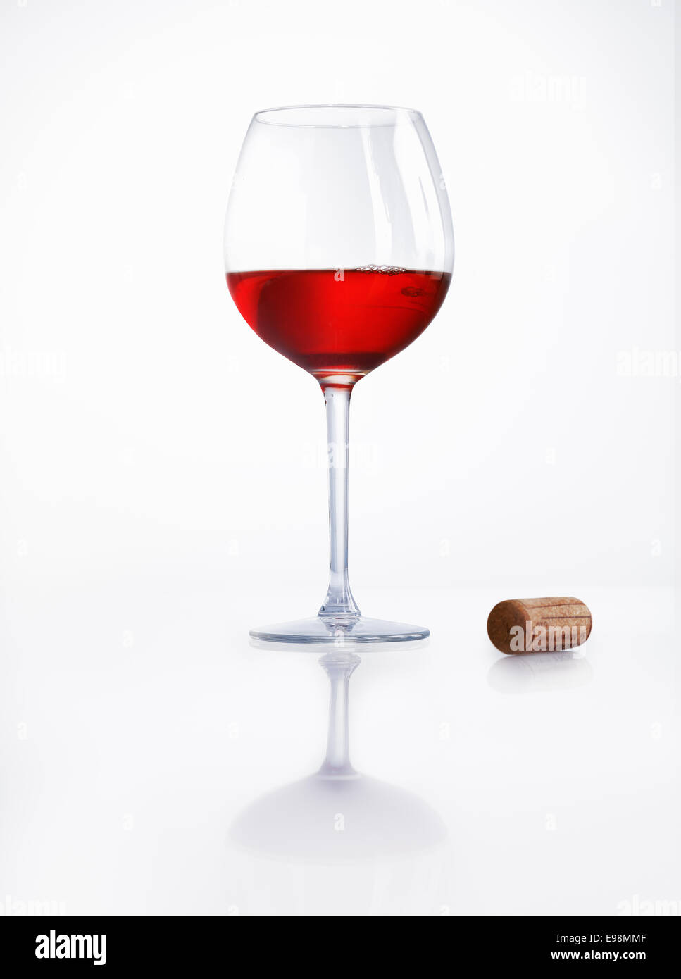 Bicchiere mezzo pieno di vino rosso su una superficie bianca riflettente con un tappo di sughero a fianco a una festa o a una celebrazione Foto Stock