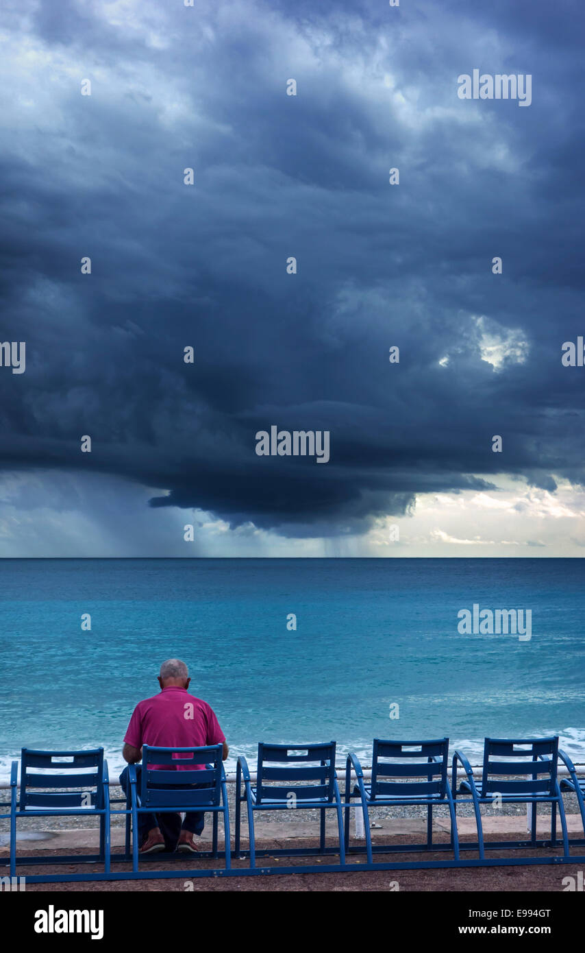 Uomo anziano seduto sul banco a dyke e guardando la scura, minacciose nuvole di tempesta sul mare Foto Stock