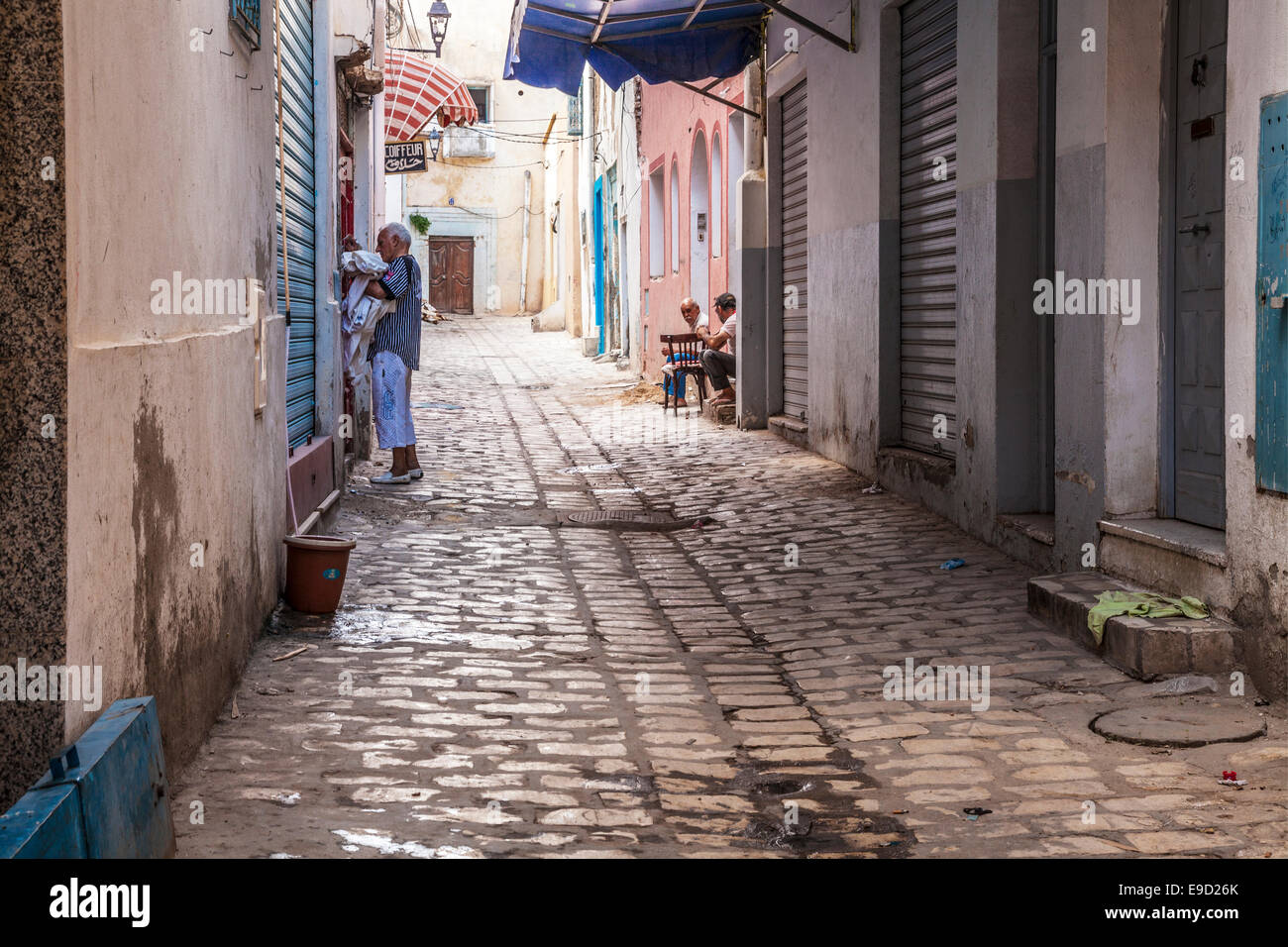 Un uomo raccoglie biancheria da una casa giù per una strada laterale nel cuore della medina a Sousse, Tunisia mentre altri due chat su una porta. Foto Stock