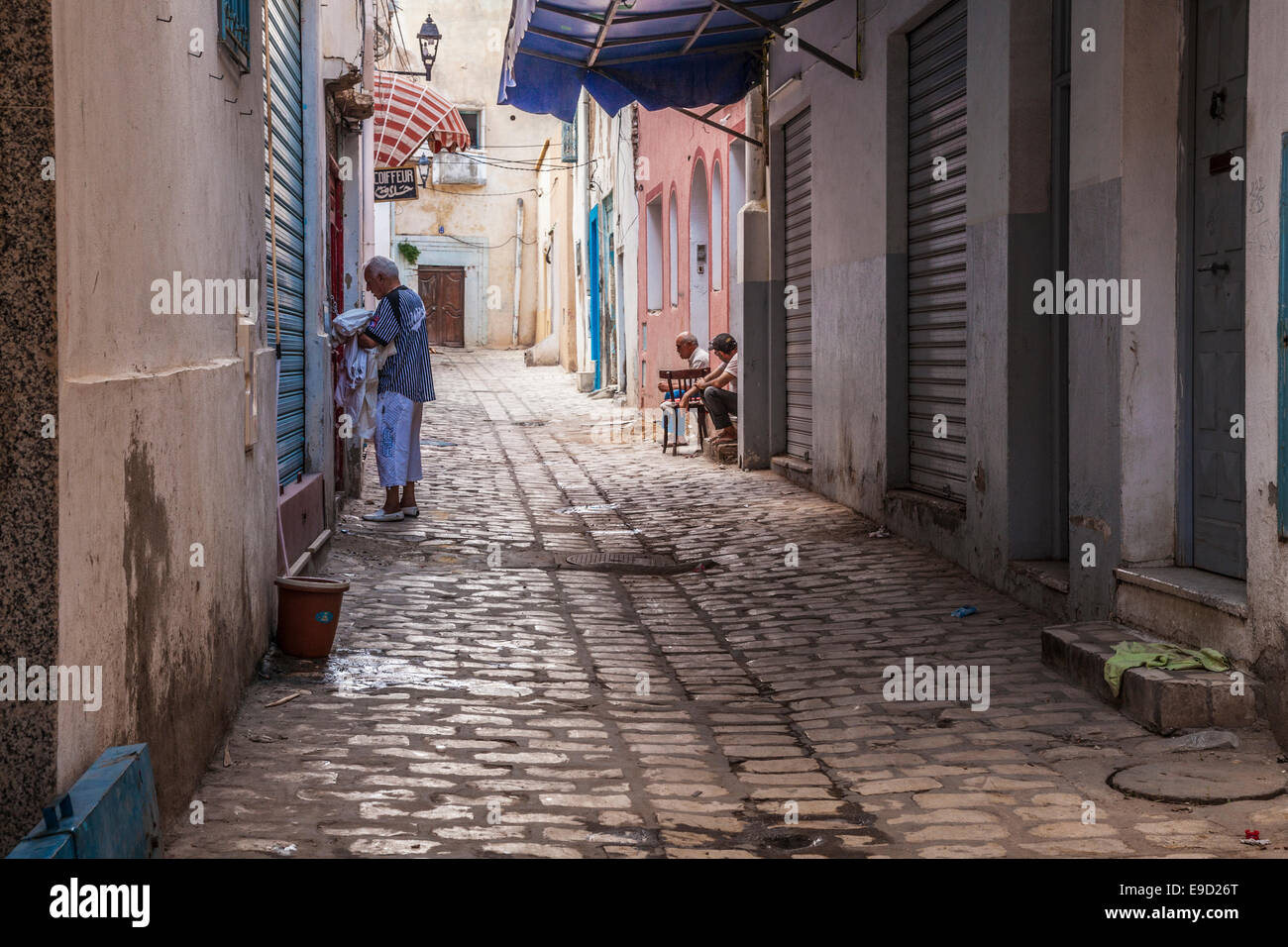 Un uomo raccoglie biancheria da una casa giù per una strada laterale nel cuore della medina a Sousse, Tunisia mentre altri due chat su una porta. Foto Stock