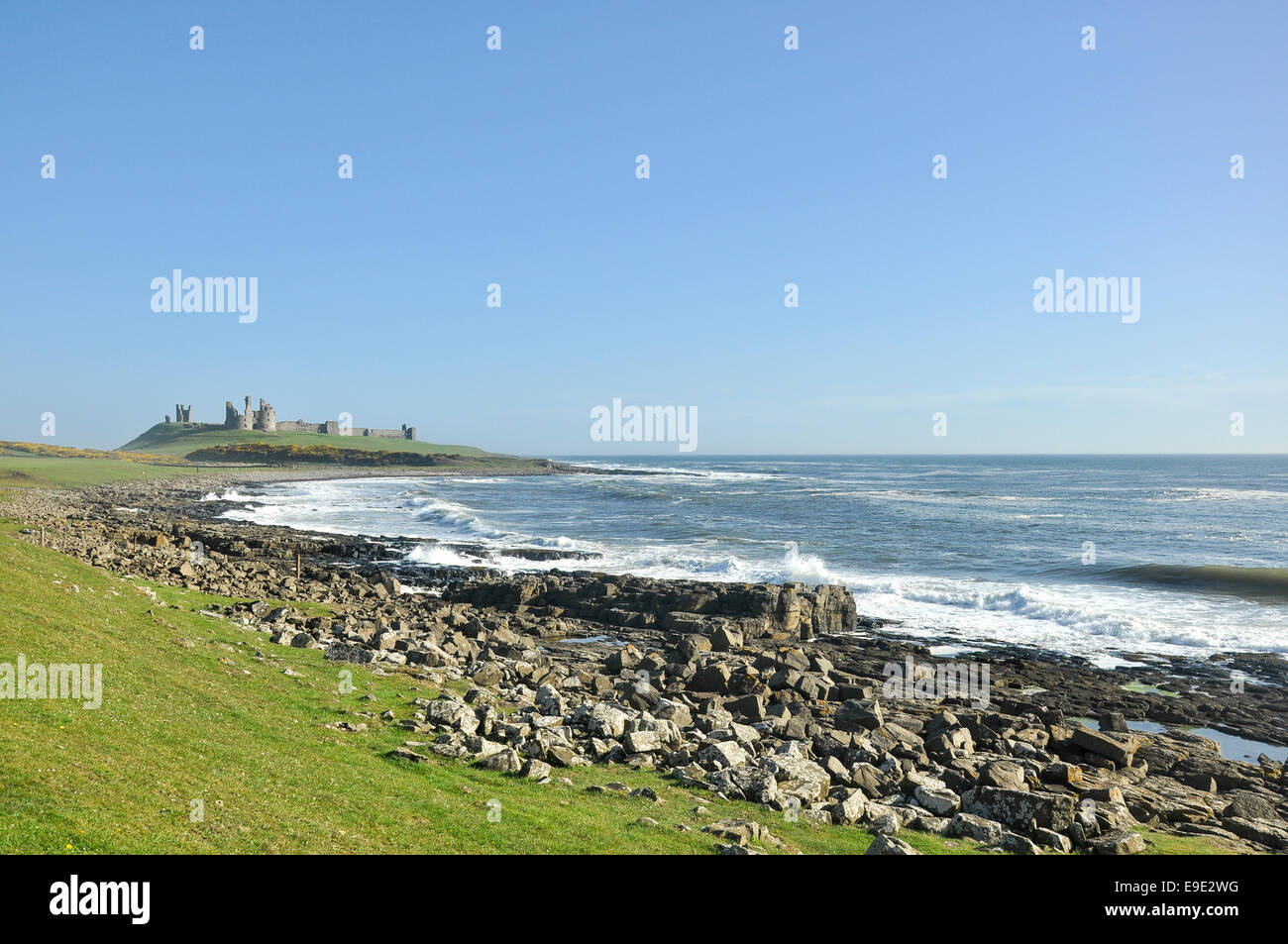 Cielo azzurro sopra il castello di Dunstanburgh in Northumberland con onde che si infrangono sulla costa rocciosa. Foto Stock