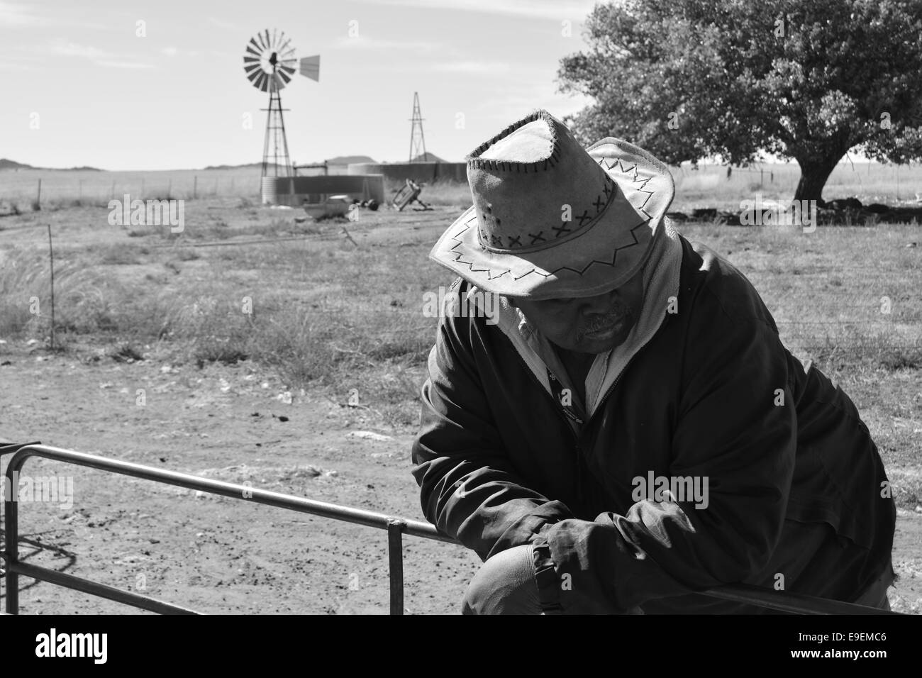 Un nero farm mano appoggiata su un recinto con un paesaggio secco in background comprendente una pompa di vento. Foto Stock