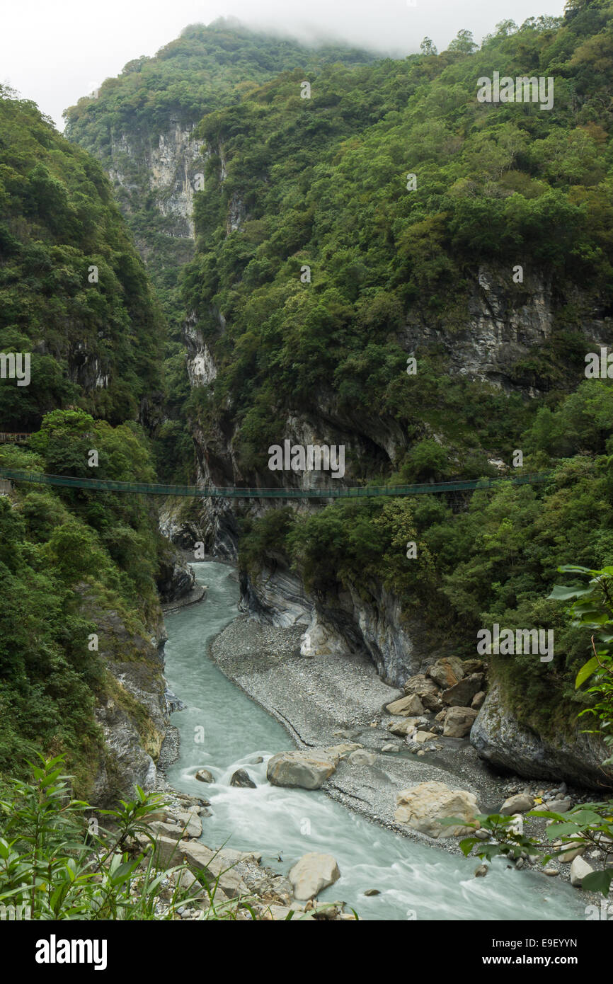 Scenario paesaggistico di lussureggianti e ripide montagne, gola profonda e una sospensione ponte su un fiume roccioso presso il Parco Nazionale di Taroko Foto Stock