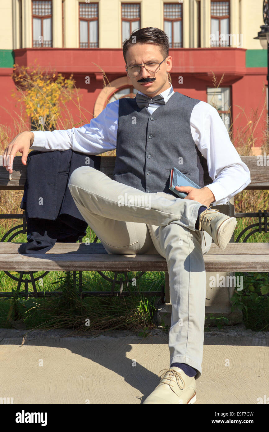 Fiducioso elegante uomo con i baffi e occhiali seduta sul banco di lavoro con un libro in mano indossando papillon in città vintage, gentiluomo su Foto Stock