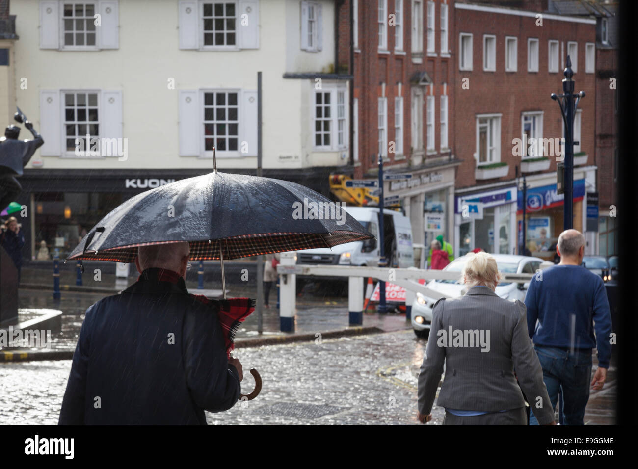 Uomo con un rosso controllare sciarpa tenendo un ombrello mentre cammina in una strada a ciottoli sotto la pioggia. Foto Stock